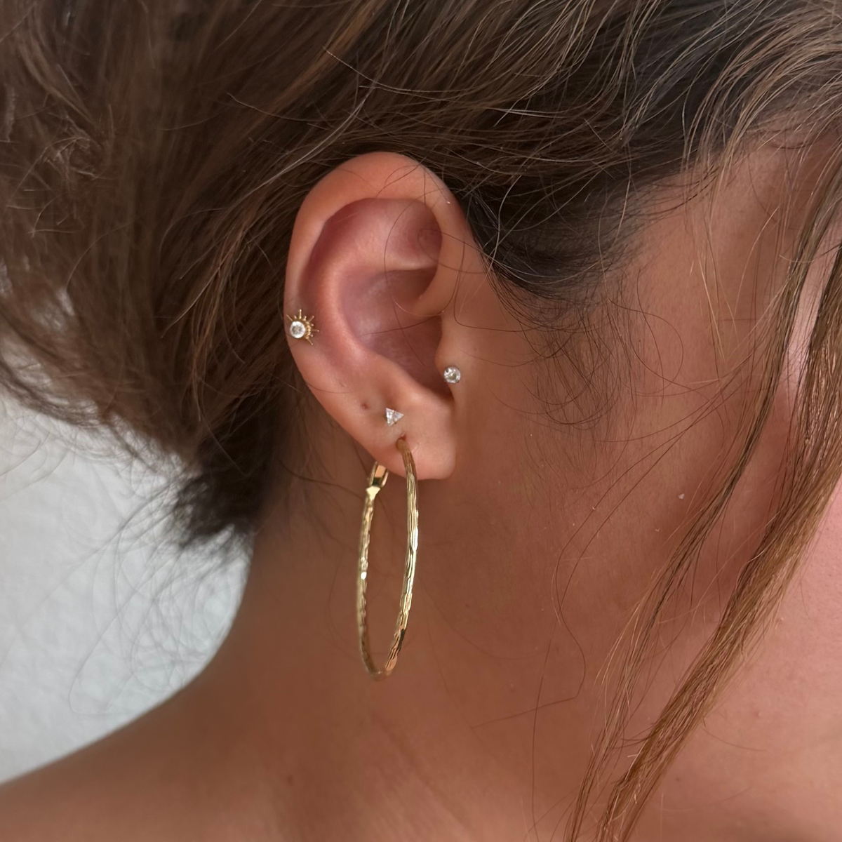 A close-up of a person's ear showcasing a stack of gold and silver stud earrings, including a large textured gold hoop, a small gold sunburst earring, and tiny silver studs.