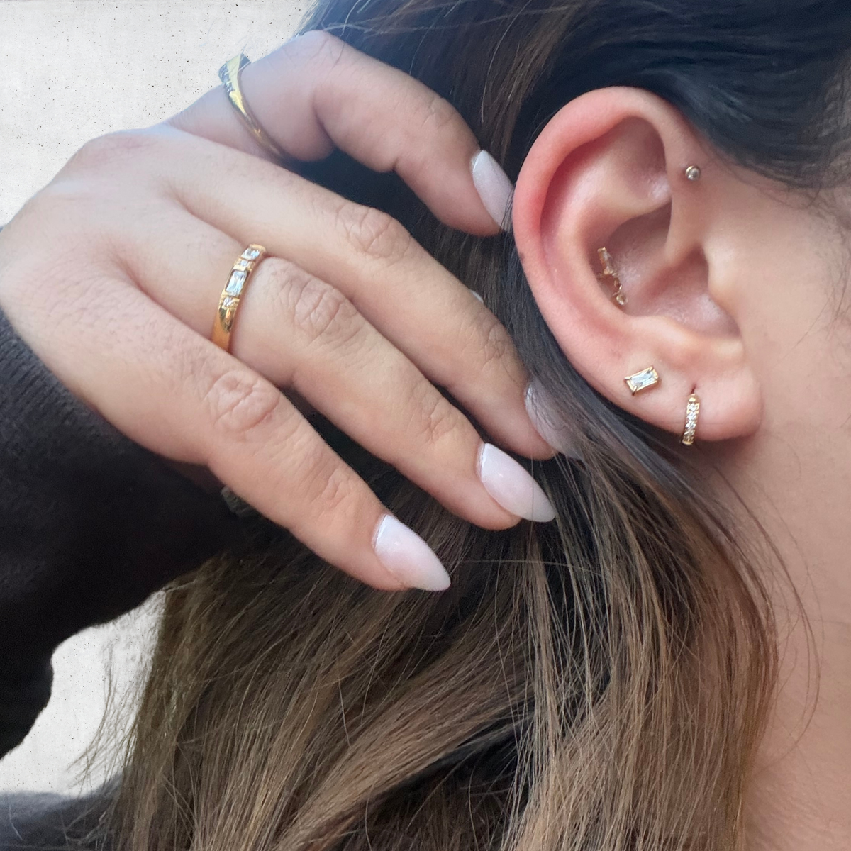 Close-up of a person's ear and hand showcasing multiple gold ear piercings and rings. The ear features several gold cartilage and lobe piercings, including a small hoop, a barbell stud, and a vertical cartilage hoop.