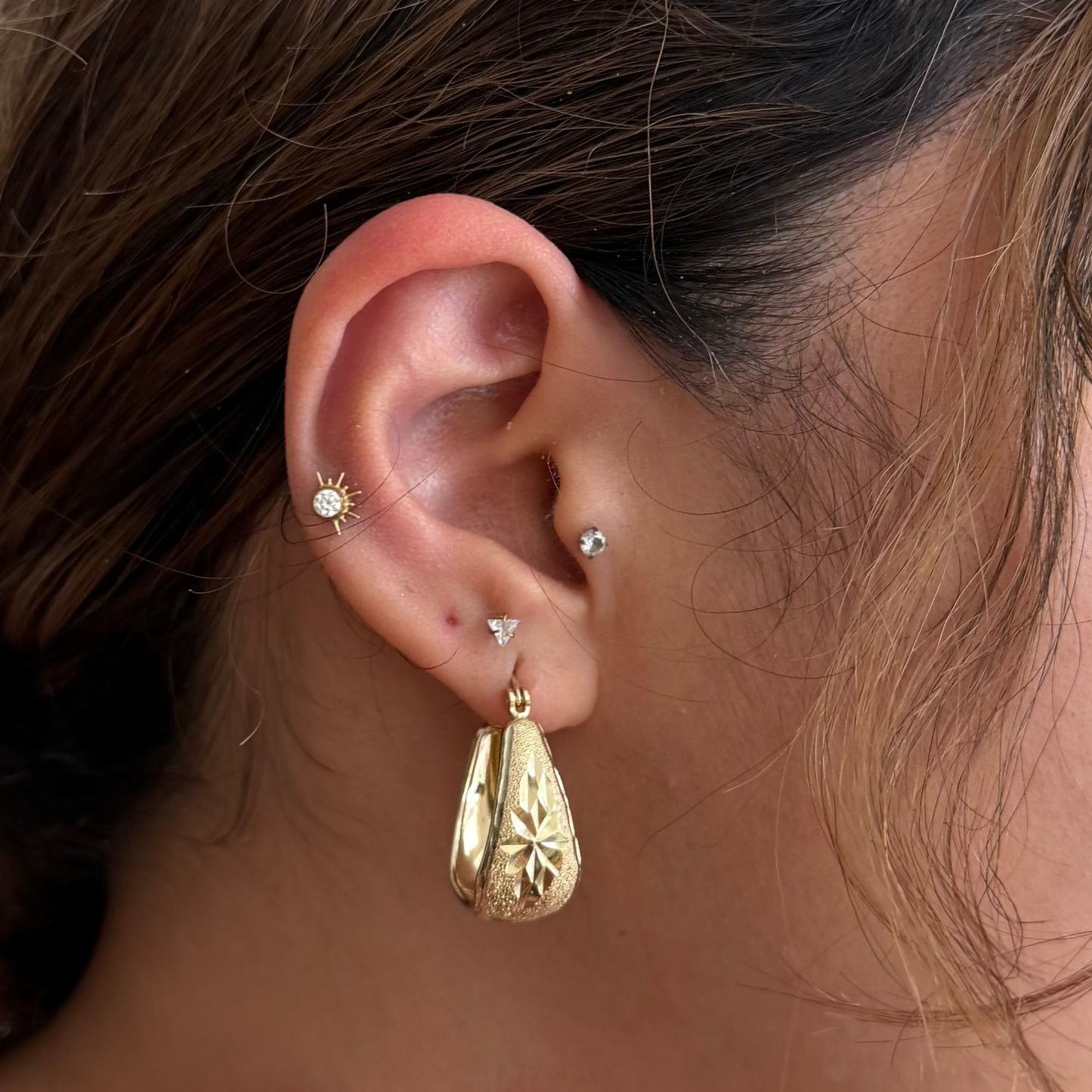 A close-up of a person's ear showcasing a gold teardrop-shaped dangle earring with an engraved floral starburst pattern, along with other small gold and crystal stud earrings.