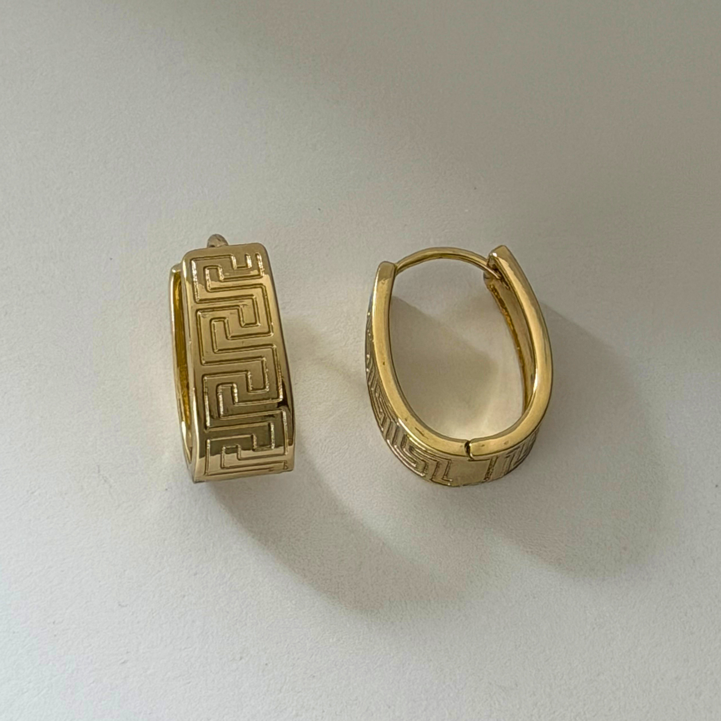 A pair of gold hoop earrings with engraved Greek key patterns, displayed on a light gray surface.