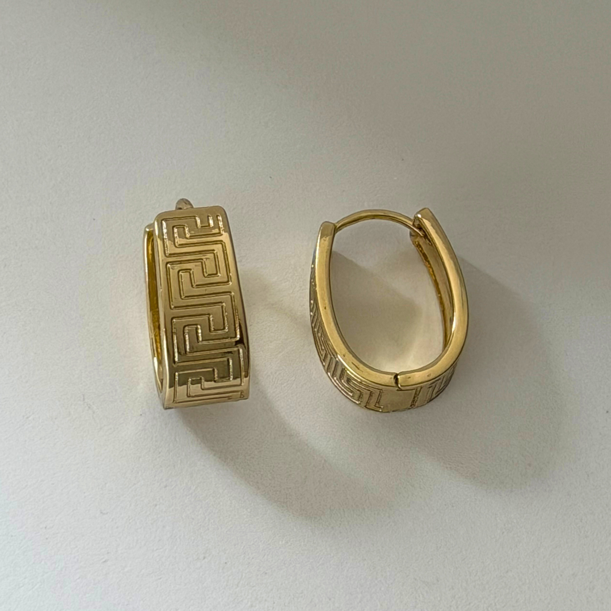 A pair of gold hoop earrings with engraved Greek key patterns, displayed on a light gray surface.