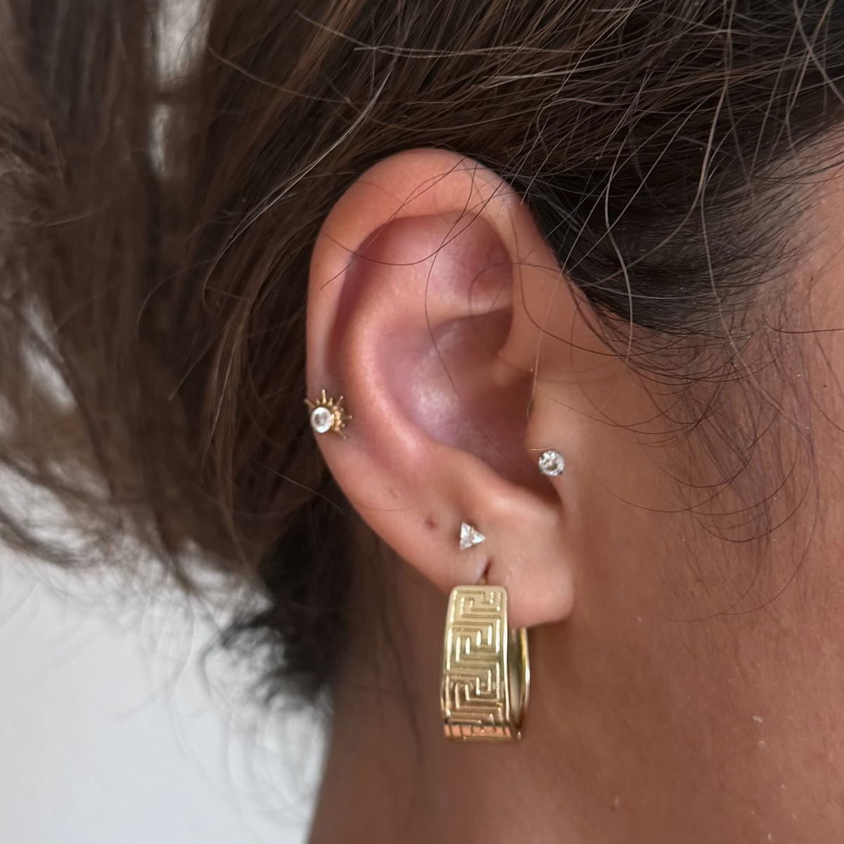 A close-up of a person's ear showcasing a stack of gold and silver earrings, including a large gold huggie hoop with a Greek key pattern, a small gold sunburst stud, a tiny silver triangle stud, and a silver ear cuff with a clear stone.