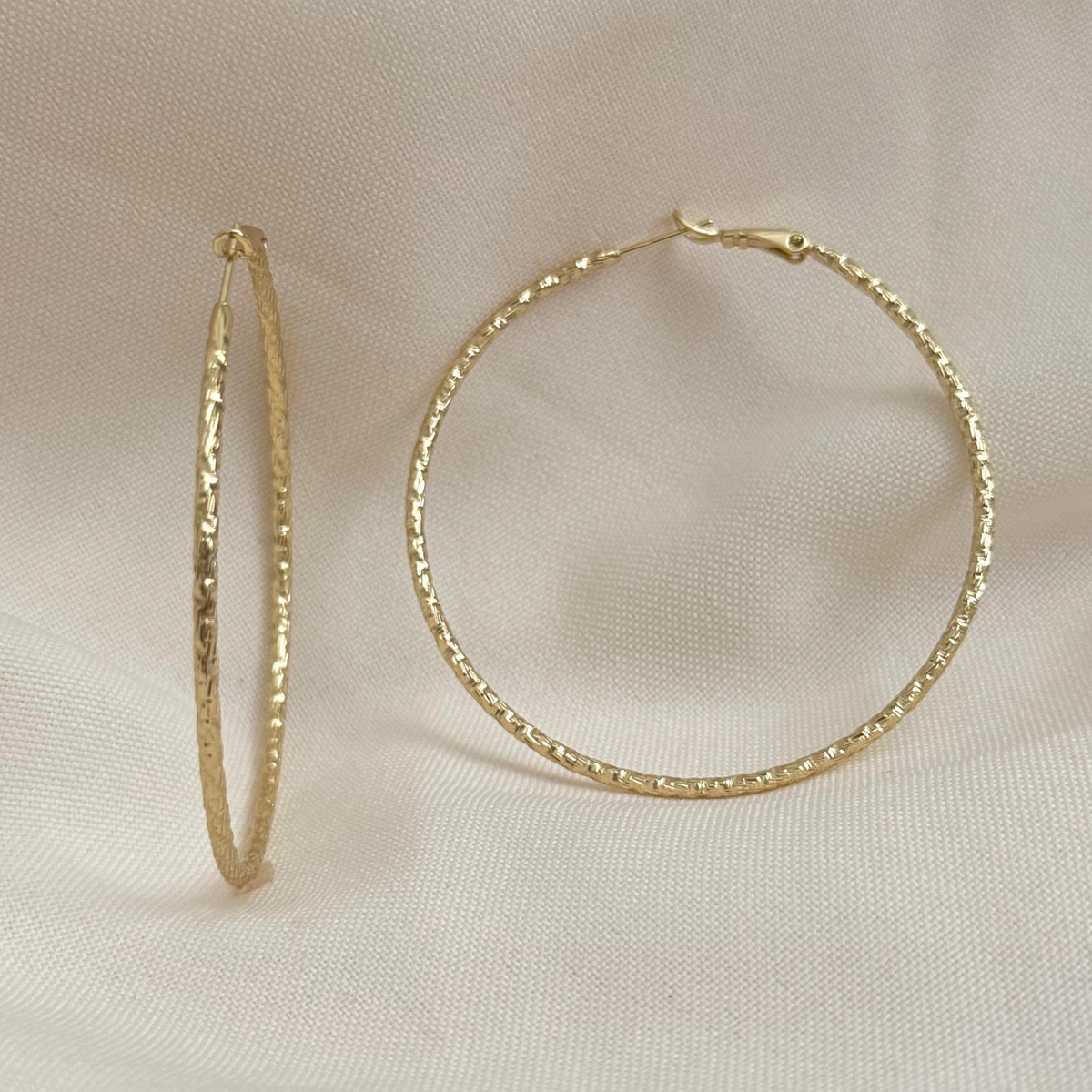 A pair of large gold plated hoop earrings with a textured, hammered finish, displayed on a soft beige fabric background.