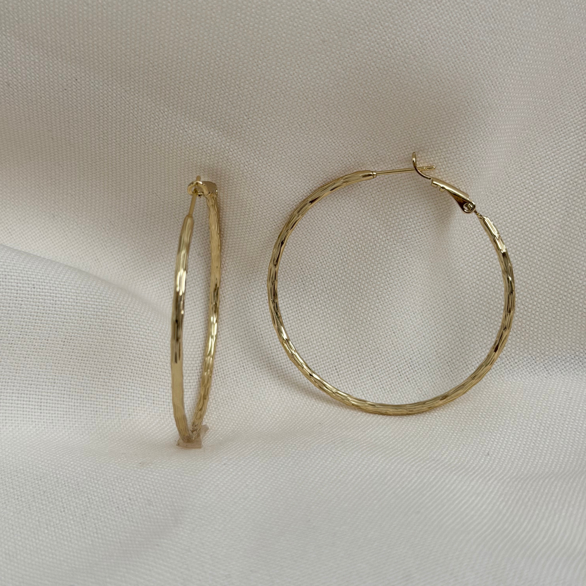 A pair of medium sized gold hoop earrings, one standing vertically and the other lying horizontally, displayed on a textured white fabric background.