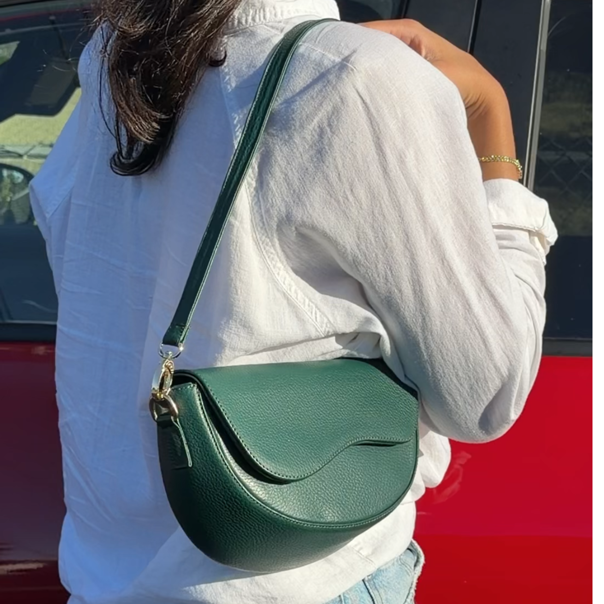 A person wearing a white shirt carries a dark green pebbled leather crescent-shaped shoulder bag with a matching strap and gold-tone hardware, photographed outdoors next to a red vehicle.