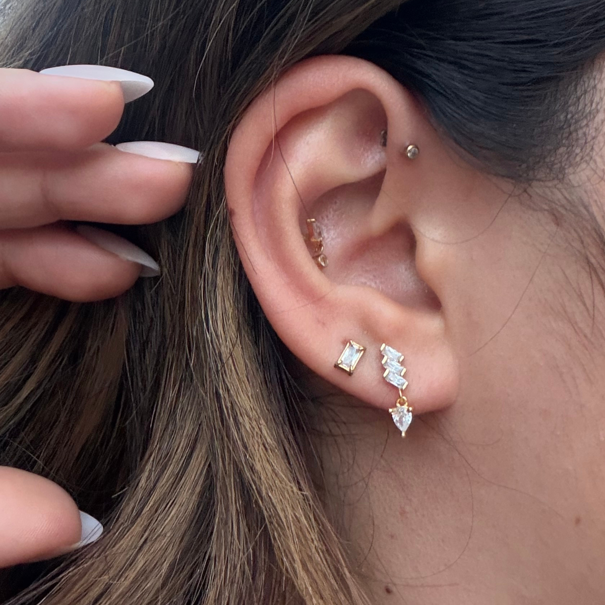 A close-up of a person's ear showcasing a multi-piece stud earring set. The set includes a small gold hoop in the cartilage, a small gold dome in the lobe, and a larger stacked cluster of gold studs with clear rectangular and marquise-cut gemstones. 