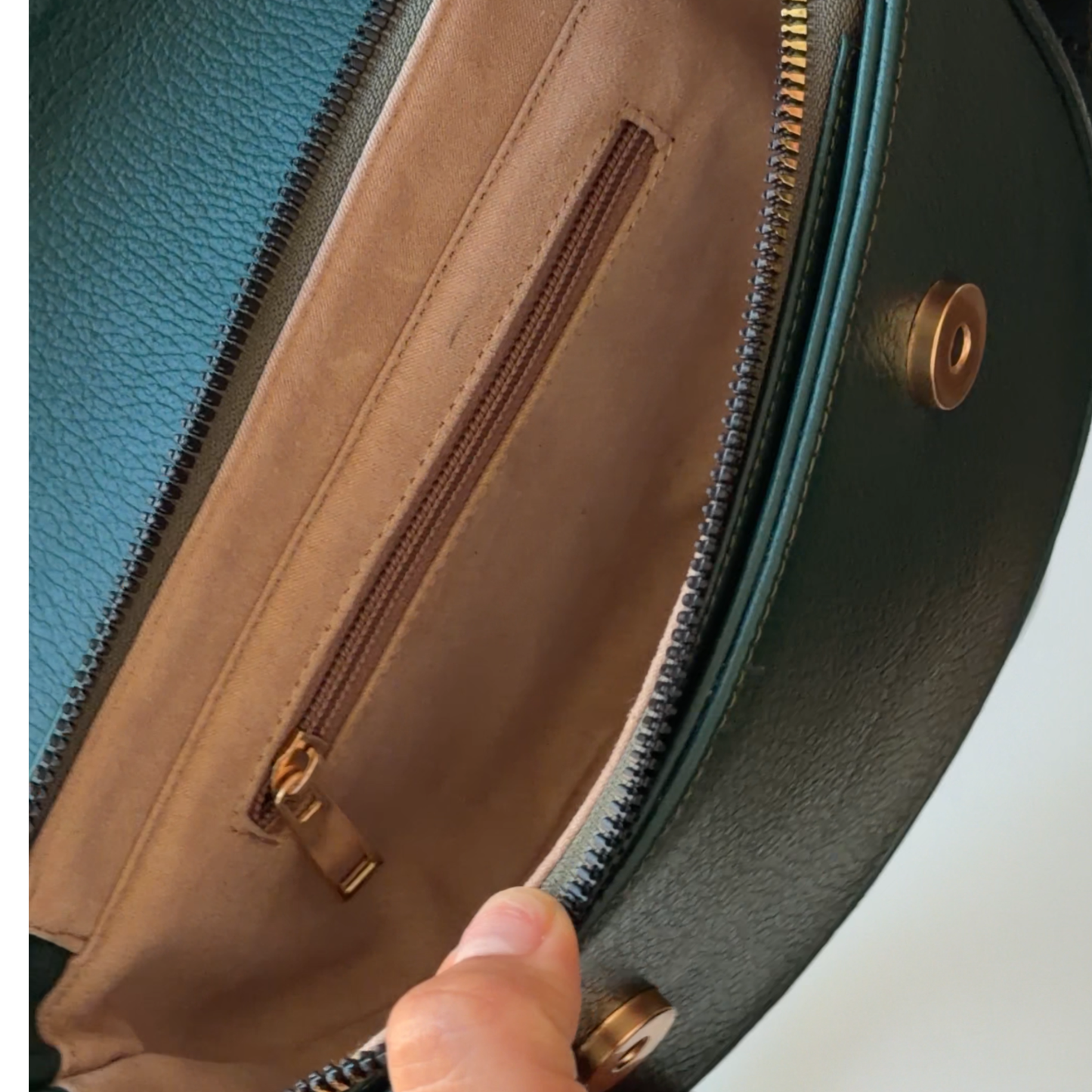 Close-up of the interior of a handbag showing a tan fabric pocket with a zipper, a dark green exterior, and a finger holding the zipper open.