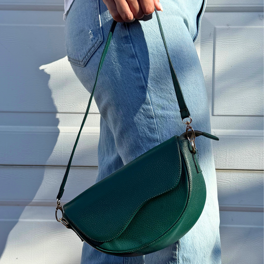 A person wearing light blue jeans holds a dark green pebbled leather crescent shoulder bag with a matching strap, set against a white paneled door.