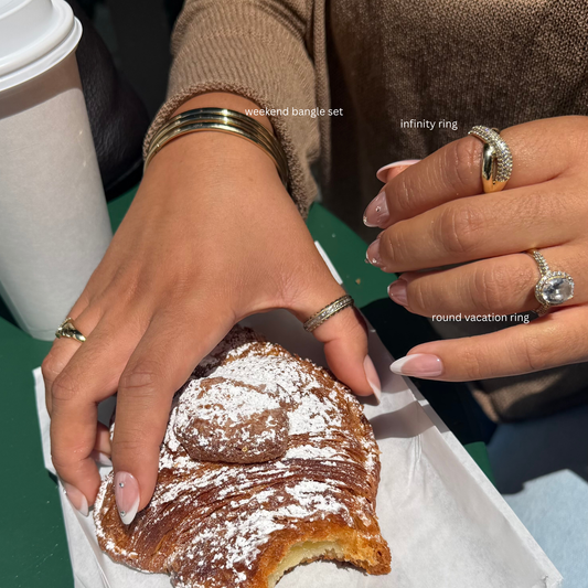 Close-up of two hands holding a powdered pastry on a tray, wearing multiple rings and bangles in a cafe setting. Text overlays identify the rings as 'infinity ring', 'round vacation ring', and 'weekend bangle set'.