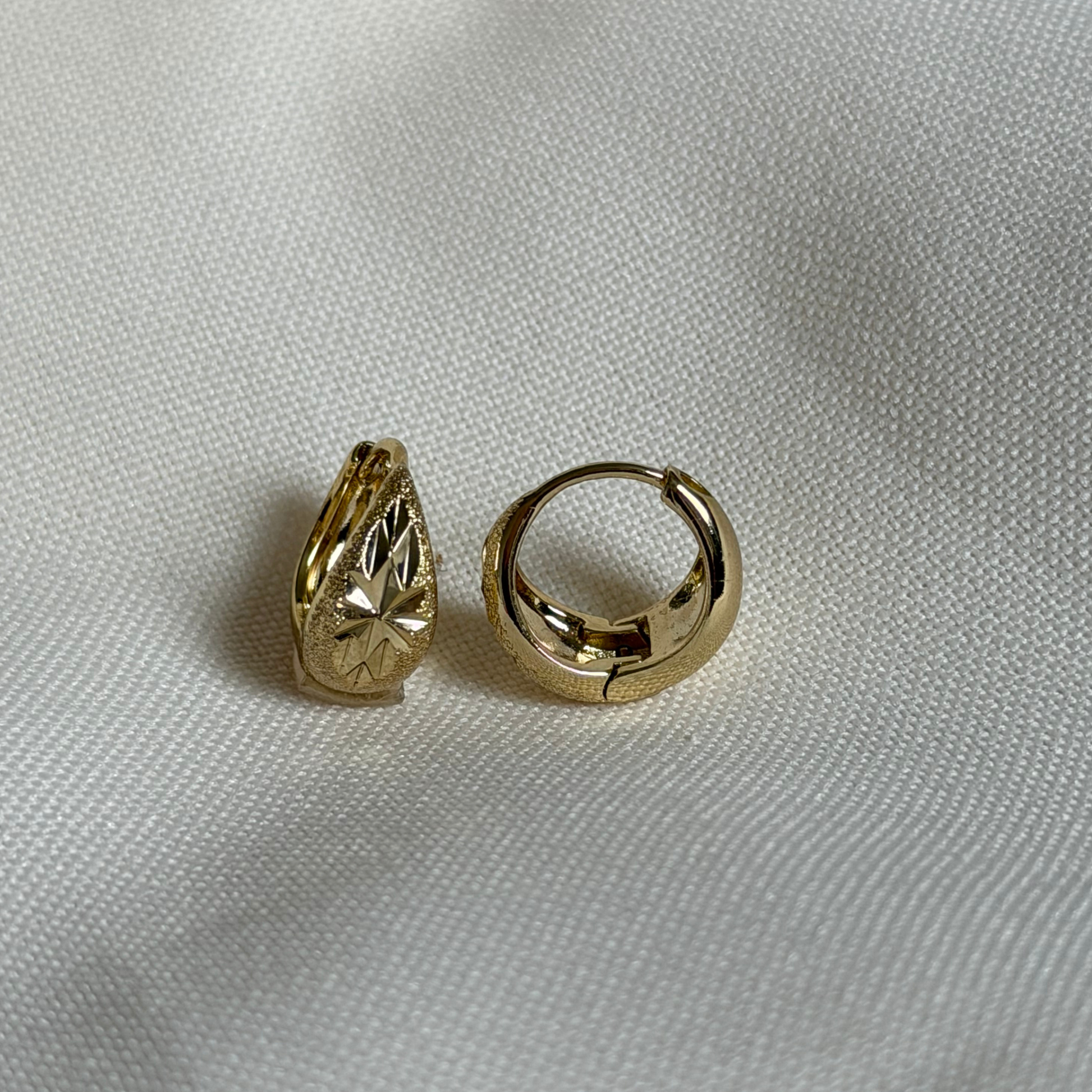 A pair of small gold hoop earrings, showing a textured floral pattern displayed on a textured white fabric background.