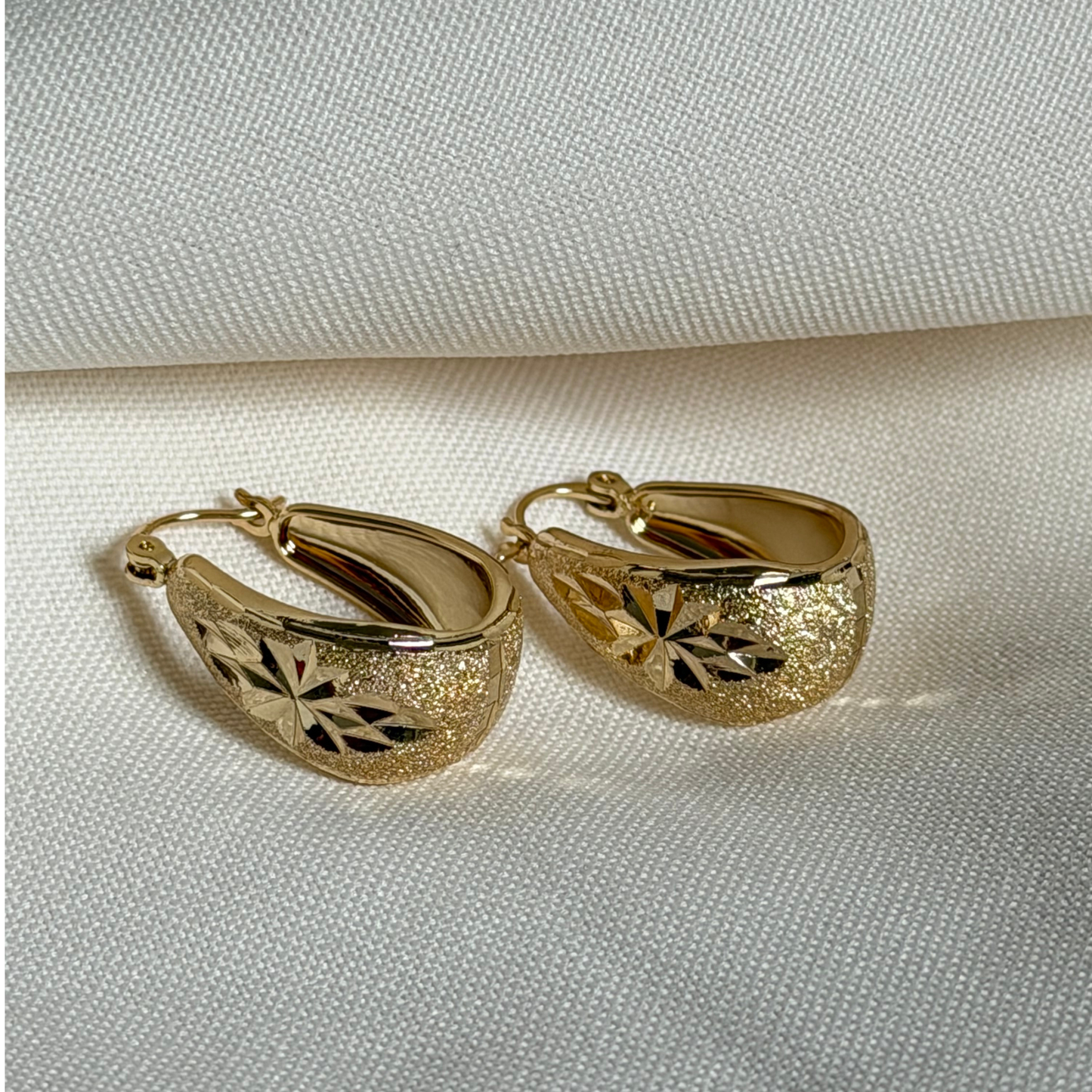 A pair of gold hoop earrings with textured glittery ends and cutout floral patterns, displayed on a textured cream fabric background.