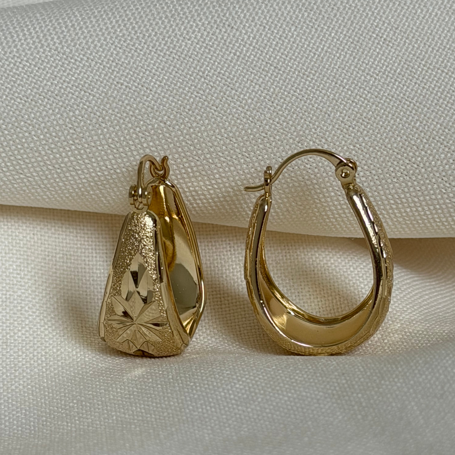 A pair of gold hoop earrings displayed on a textured cream fabric background. One earring is shown from the front, featuring a textured, starburst-patterned teardrop-shaped face, while the other is shown from the side, displaying a smooth, polished crescent shape.