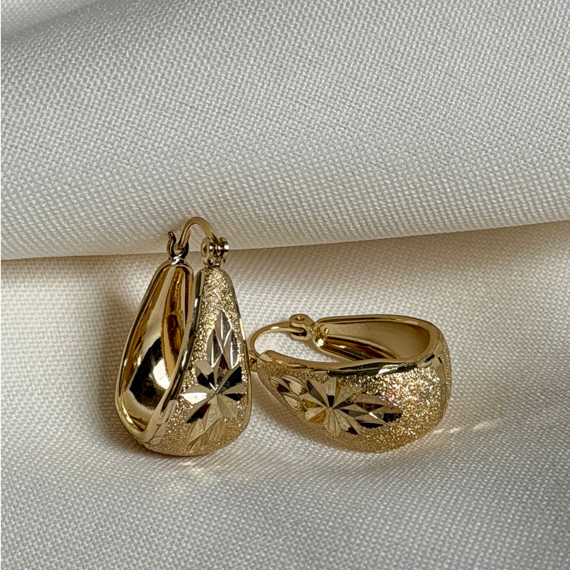 A pair of gold-toned hoop earrings with textured, starburst-patterned metal, displayed on a textured off-white fabric background.