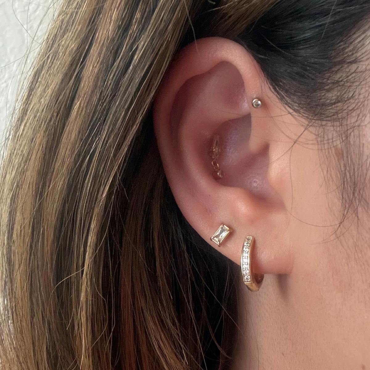 Close-up of a person's ear showcasing multiple gold piercings, including a hoop stud in the lobe, a small rectangular stud, a tiny cartilage stud, and a cartilage piercing, with brown hair framing the side of the face.