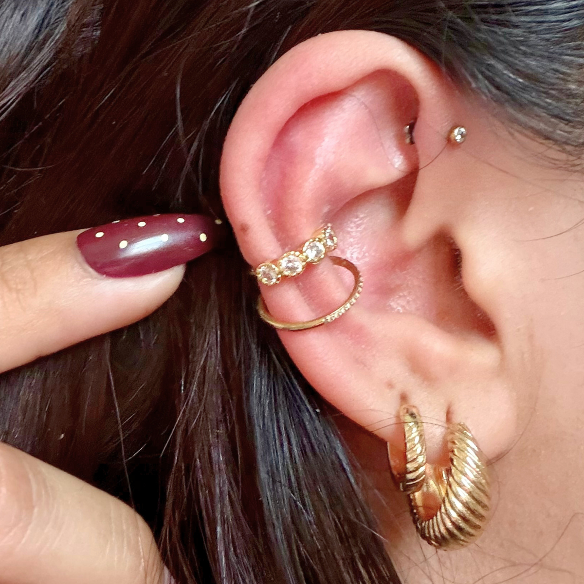 Close-up of an ear with gold hoop earrings and a ring, with a hand adjusting the jewelry.